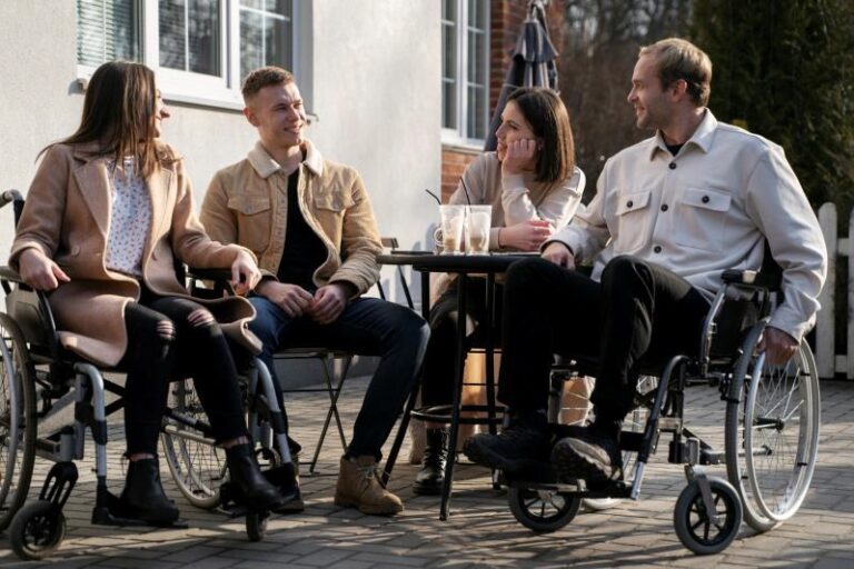 Four people outdoors, having a conversation around a round table. Two of them are in wheelchairs, and they are all smiling.