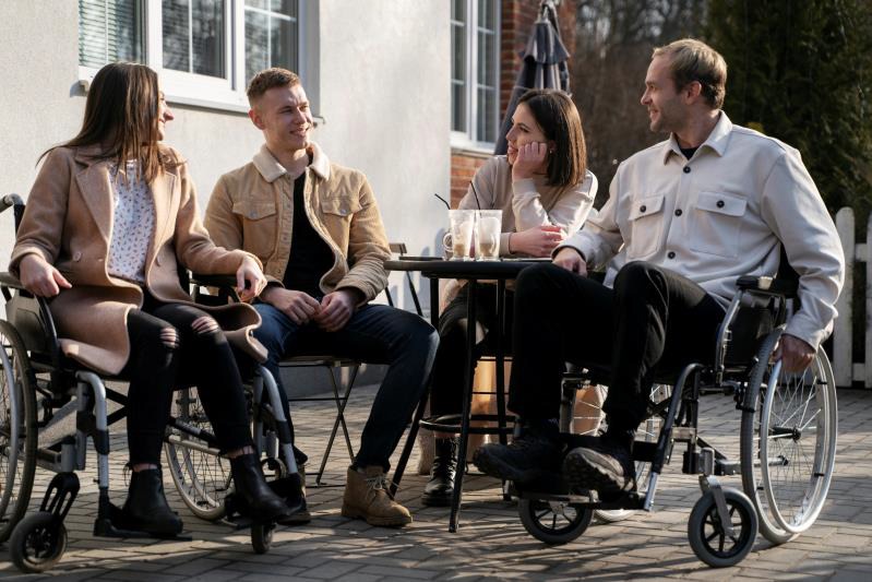 Four people outdoors, having a conversation around a round table. Two of them are in wheelchairs, and they are all smiling.